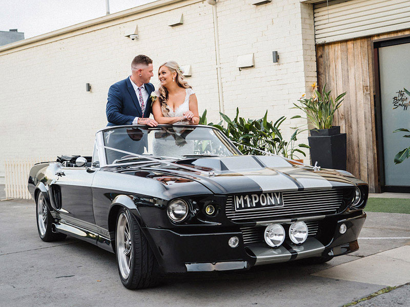 Bride and Groom standing in a black Mustang.