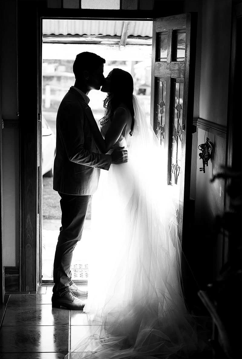 Bride and groom kissing in the doorway with gown illuminated by light.