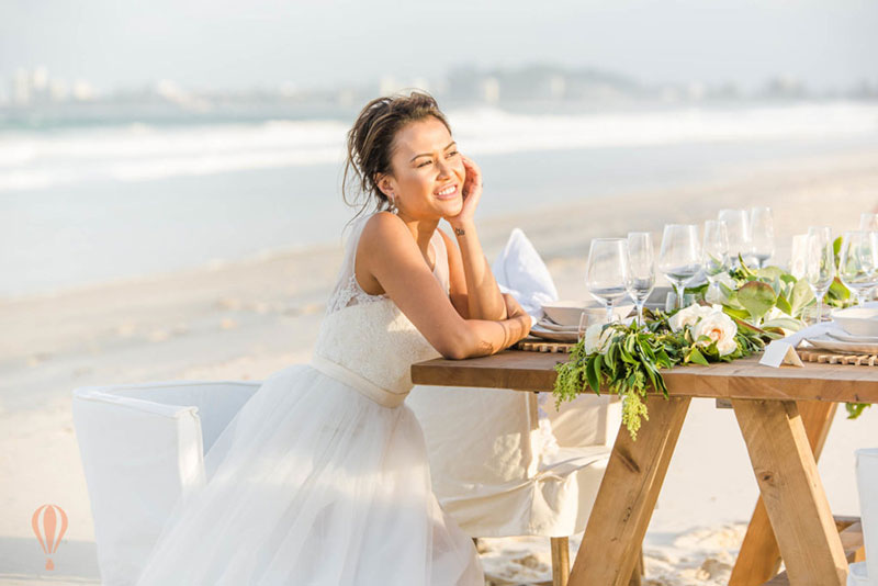 Happy bride sits smiling at reception table set up on beach.