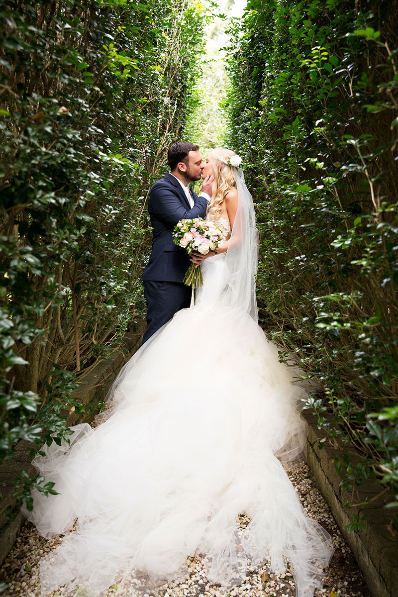 Bride and Groom kissing between tall hedges.