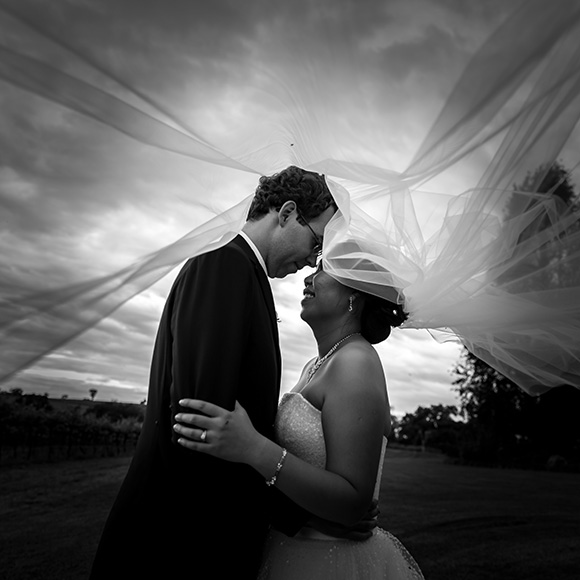 Black and white photo of Margaret and Michael holding eachother under a cloudy sky with Margaret's veil enveloping them.