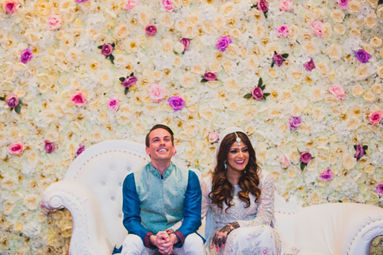 Multicultural Bride and Groom sitting together on a white couch in front of a flower wall.