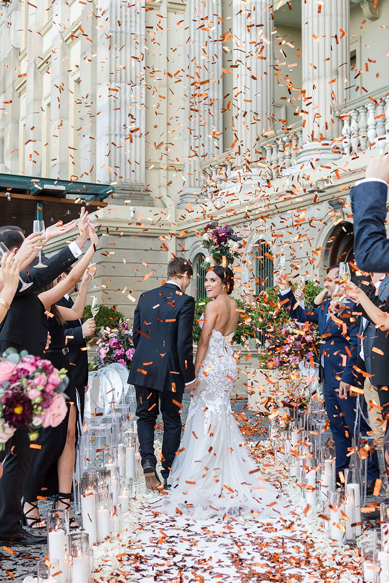 Smiling Bride and Groom walk between guests and bursts of colourful confetti.